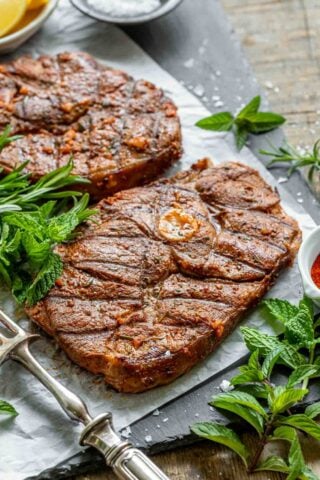 two pieces of lamb steak on a grey surface with fresh greens scattered around them