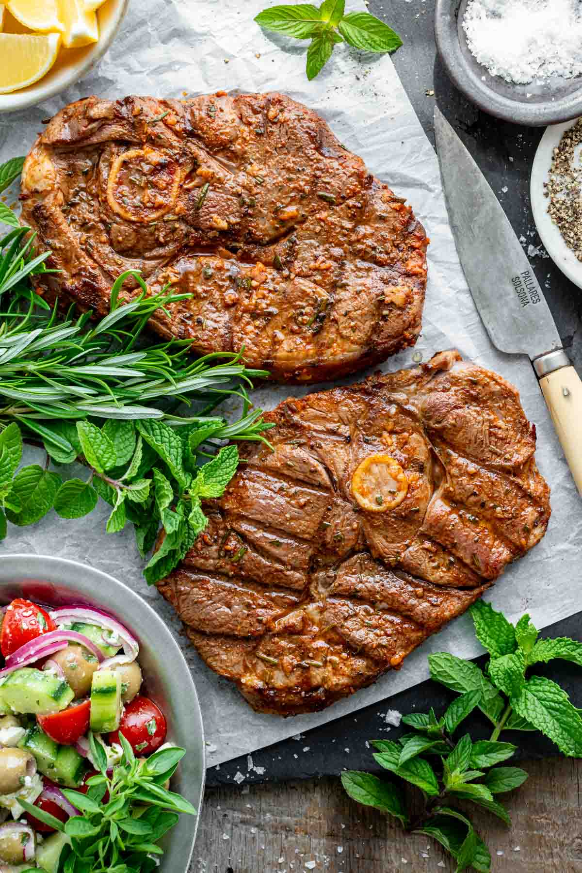 Two lamb legs on a grey surface with a mediterranean cucumber salad in the bottom left corner