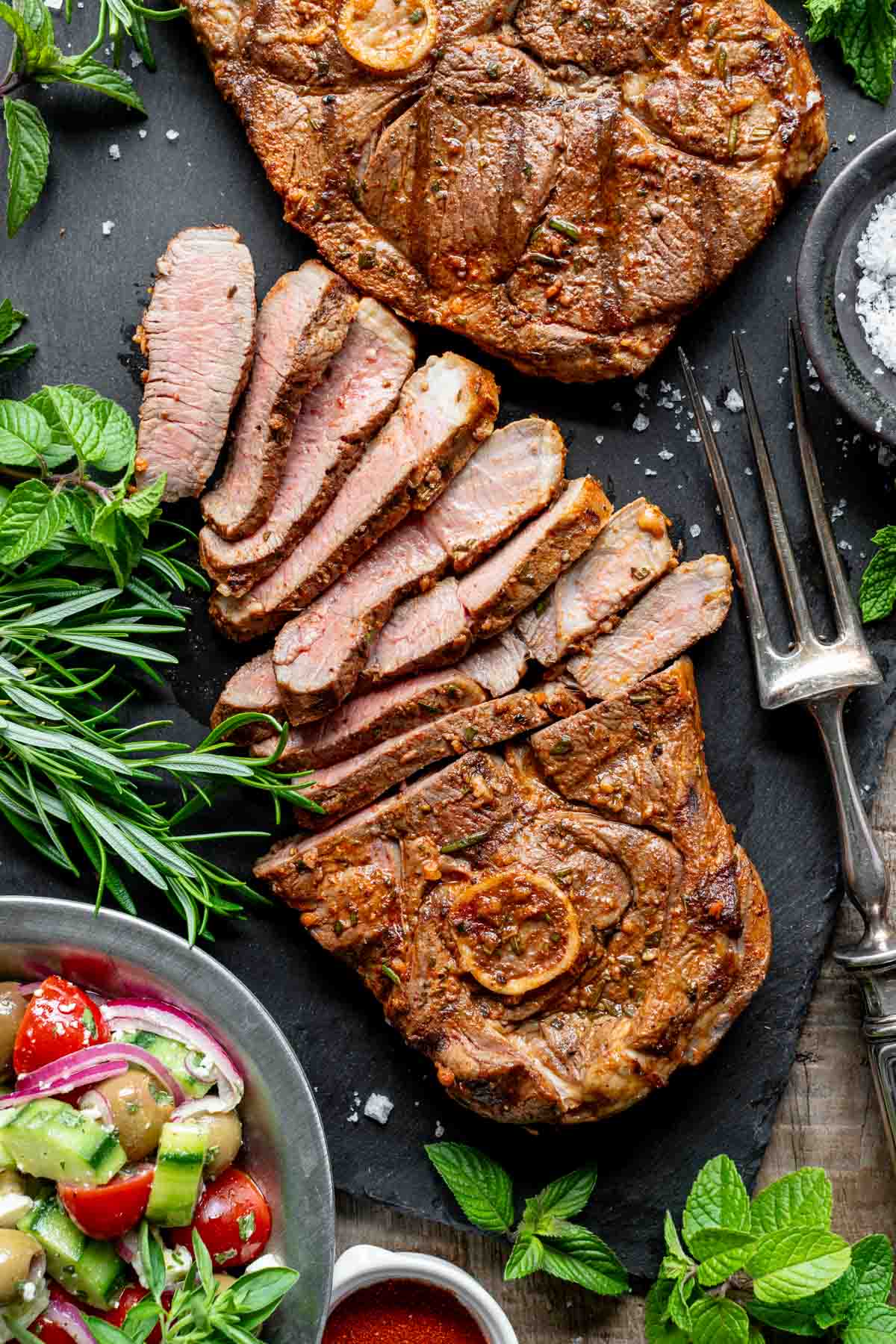 lamb steak on a charcoal background with half of it sliced into strips and a bowl of greens and salad beside it in the lower left corner
