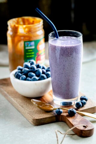high protein blueberry peanut butter smoothie in glass cup with a bowl of blueberries and peanut butter jar behind