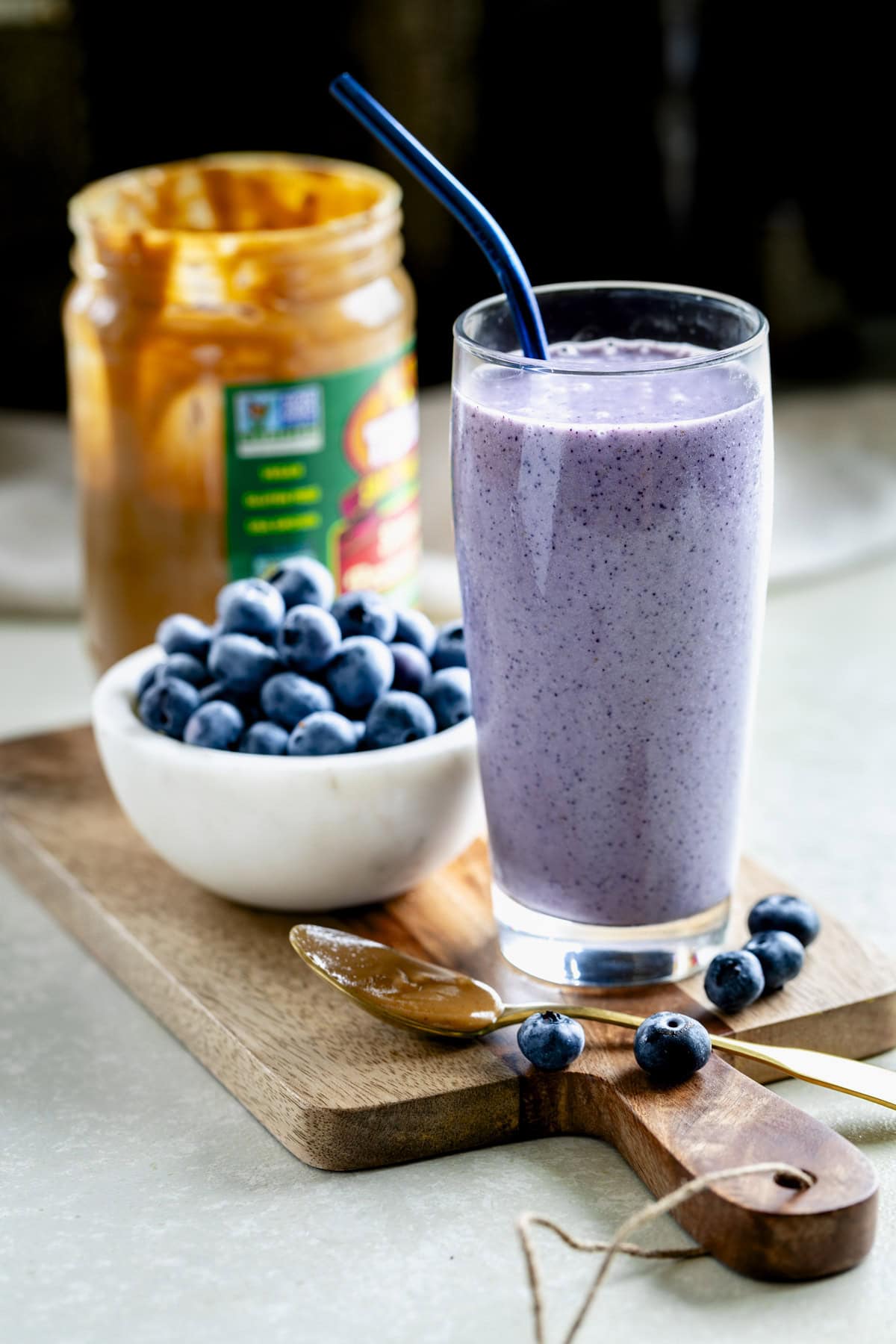 high protein blueberry peanut butter smoothie in glass cup with a bowl of blueberries and peanut butter jar behind
