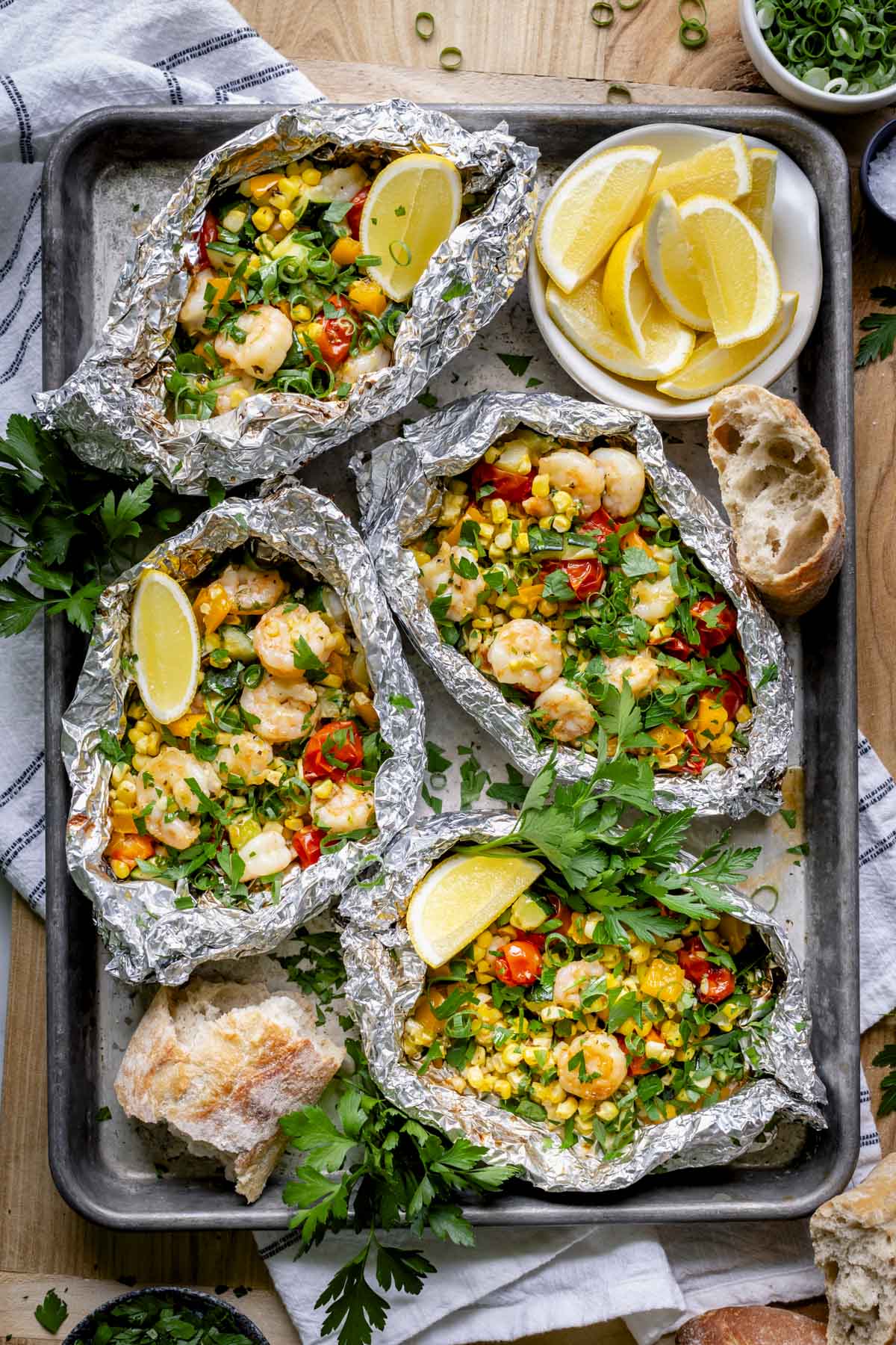 four packets of grilled shrimp and veggies in foil packets on a baking sheet with crusty bread and lemon wedges