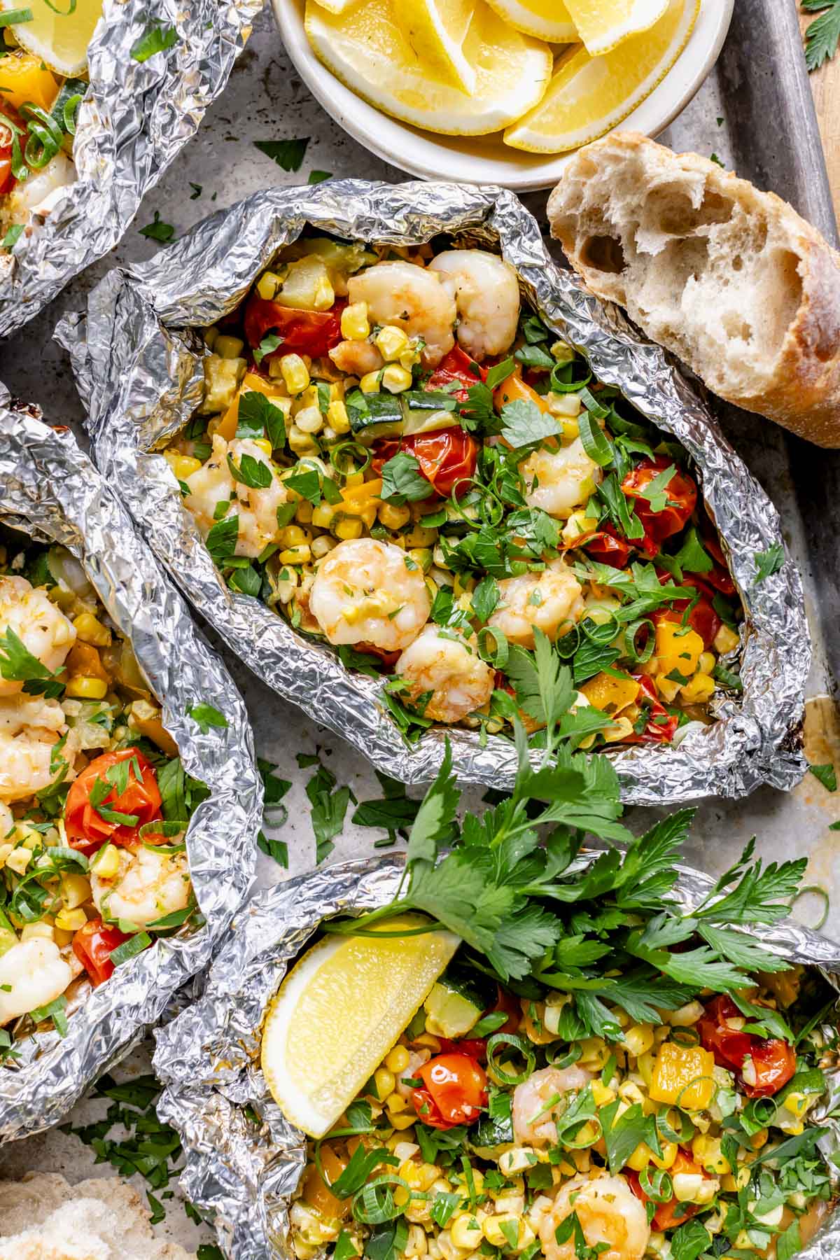 a closeup photo of a couple of the foil packets full of shrimp, vegetables, and parsley garnish beside a crusty piece of bread