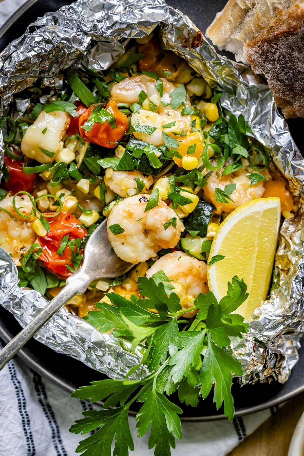 a bowl made of foil full of grilled shrimp and veggies garnished with parsley and lemon juice